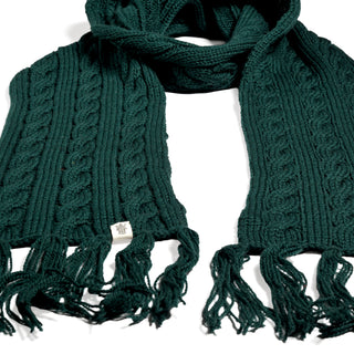 The Hand Knit Plush Scarf, crafted from soft Merino wool in dark green, features braided cable patterns and fringe at both ends. Laid flat on a white background, it displays a small rectangular tag attached near one end.