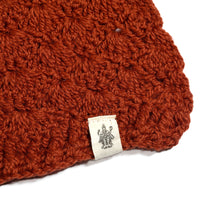 A close-up of a textured, rust-colored bobble stitch fabric crafted for the Hand Crochet Veil Scarf, showcasing a small white tag with a black line drawing of a figure attached to the edge of the hand-crocheted Merino wool scarf.