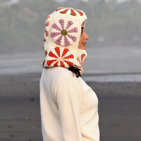 A person stands in profile on a beach, wearing the Hand Crochet Petal Hood, a white hooded garment featuring a red and purple floral crochet pattern and a cozy sherpa fleece lining. The background is blurred, with dark sand and some greenery visible.