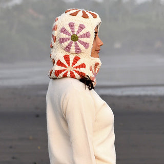 A person stands in profile on a beach, wearing the Hand Crochet Petal Hood, a white hooded garment featuring a red and purple floral crochet pattern and a cozy sherpa fleece lining. The background is blurred, with dark sand and some greenery visible.
