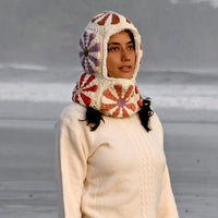 A person wearing a cream sweater with sherpa fleece lining and the Hand Crochet Petal Hood—a colorful crochet hat adorned with flower patterns—stands on a foggy beach, looking slightly upward. The background shows sand and a blurry shoreline.