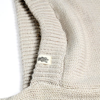 A close-up of the Hand Loom Nomad Hood Scarf in beige showcases its textured knit and precise stitching, with a small white tag displaying a black crocodile logo sewn into the seam. The image emphasizes the intricate craftsmanship and cozy appeal of this hooded scarf.
