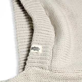 A close-up of the Hand Loom Nomad Hood Scarf in beige showcases its textured knit and precise stitching, with a small white tag displaying a black crocodile logo sewn into the seam. The image emphasizes the intricate craftsmanship and cozy appeal of this hooded scarf.