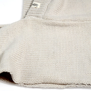A close-up of the beige Hand Loom Nomad Hood Scarf showcases its intricate knitted texture and stitching patterns, with a small white and black tag sewn near the top edge. The detailed craftsmanship highlights what makes this lambswool scoodie both cozy and stylish.