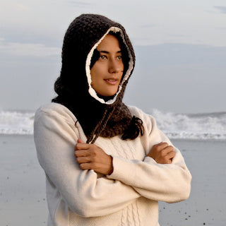 Wearing the Hand Crochet Cloud Hood and a cream-colored sweater, a person stands on the beach with arms crossed, framed by waves and a cloudy sky in the background.