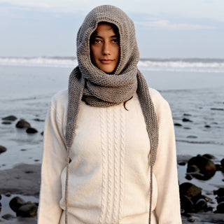 A person stands on a rocky beach wearing a cream-colored textured sweater and the Anna Hood, a chunky gray wool winter accessory handmade in Nepal; the ocean and cloudy sky are in the background.