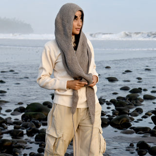 Wearing the Anna Hood beige sweater, tan cargo pants, and a gray knit scarf—an ideal winter accessory—a person stands on a rocky beach with waves in the background.