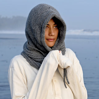 A person stands near the ocean wearing the Hand Knit Halo Reverse Hood in a thick gray lambswool, styled as both hood and scarf. Paired with a white long-sleeve shirt, they look slightly to the side with hands partially hidden in their sleeves. Behind them, waves roll along a misty shoreline.