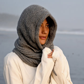A person stands outdoors by the water on a foggy day, looking ahead with their hands partly covered by their sleeves, wearing the thick gray Hand Knit Halo Reverse Hood over a white sweater.