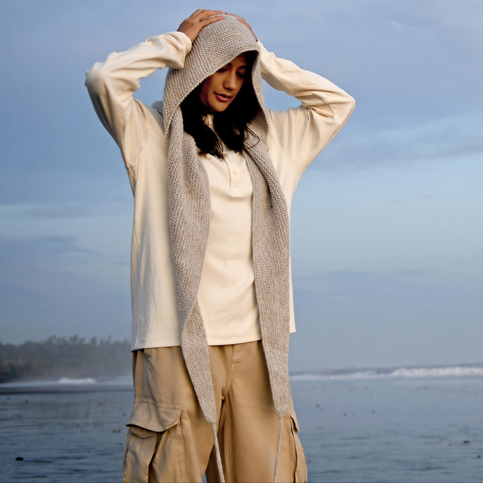 Wearing the Hand Knit Anna Hood—a light-colored, sculpted hood with an integrated scarf—a person stands on a beach with their hands on their head, beige cargo pants complementing the look. Cloudy skies and the ocean form the backdrop.