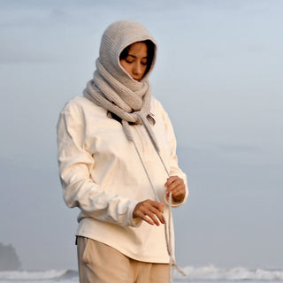 Wearing the Anna Hood, a person stands outdoors in the mist, looking down and holding a strand of yarn. With its cozy wool hood and thick knit scarf, this cream-colored accessory stands out against the foggy backdrop of blurred trees and water—perfect for staying warm all winter.