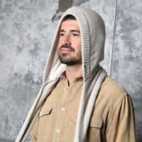 A man with a beard wears a tan button-up shirt and the Hand Loom Nomad Hood Scarf, a beige hand-knit hooded accessory styled as a cozy scoodie. He stands before a textured gray wall, gazing slightly upward and to the side.