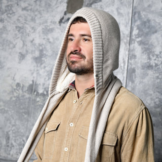 A man with a beard wears a tan button-up shirt and the Hand Loom Nomad Hood Scarf, a beige hand-knit hooded accessory styled as a cozy scoodie. He stands before a textured gray wall, gazing slightly upward and to the side.