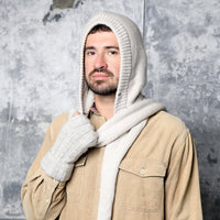 A man wearing a beige button-up shirt and the Hand Loom Nomad Hood Scarf with matching gloves stands in front of a textured gray wall, looking at the camera with a neutral expression.