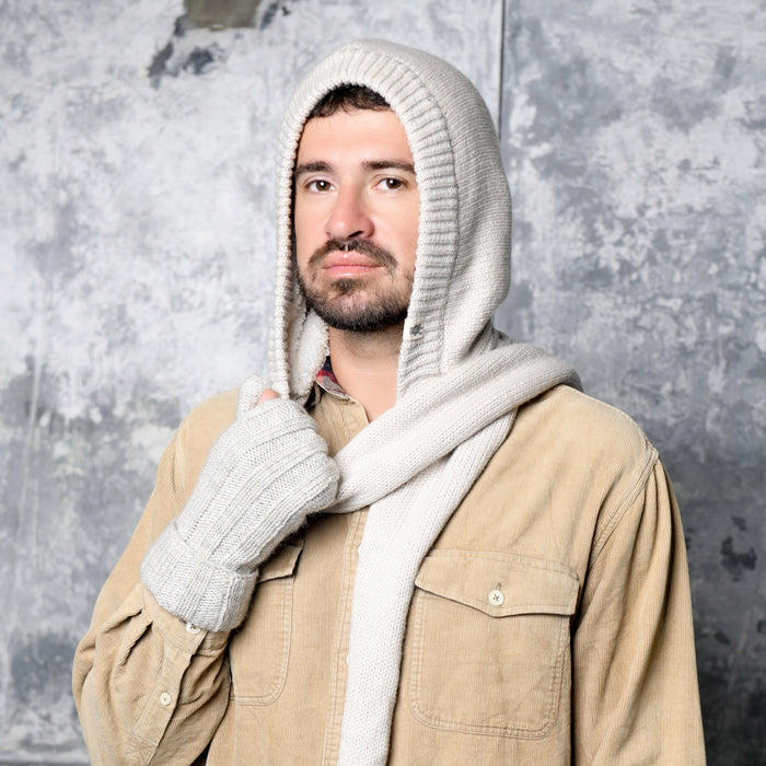 A man wearing a beige button-up shirt and the Hand Loom Nomad Hood Scarf with matching gloves stands in front of a textured gray wall, looking at the camera with a neutral expression.