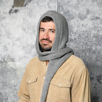 A man with a beard wears a tan button-up shirt and the Hand Loom Nomad Hood Scarf, a gray hand-knit hooded scarf wrapped around his head and neck, standing against a textured gray background.