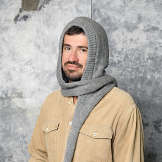 A man with a beard wears a tan button-up shirt and the Hand Loom Nomad Hood Scarf, a gray hand-knit hooded scarf wrapped around his head and neck, standing against a textured gray background.