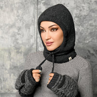 A person wearing the Hand Knit Cozy Hood and matching fingerless gloves in gray looks to the side against a gray textured background, holding the drawstrings of the hooded balaclava.