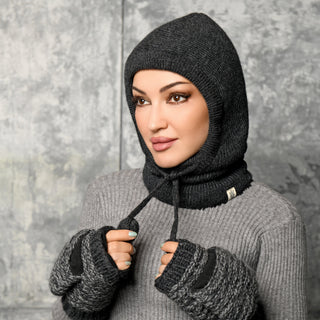 A person wearing the Hand Knit Cozy Hood and matching fingerless gloves in gray looks to the side against a gray textured background, holding the drawstrings of the hooded balaclava.