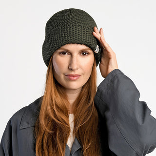 A person with long red hair wears the Baltic Hat, a dark green winter hat handmade in Nepal, along with a gray oversized jacket. They look directly at the camera and touch the side of the hat with one hand.
