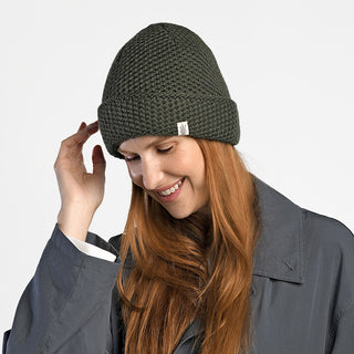 A smiling woman with long red hair wears the handmade Baltic Hat in dark green wool, paired with a gray coat. She looks down, touching the side of her hat against a plain white background.