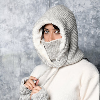 Wearing the Hand Knit Glide Hood in light gray—crafted from soft lambswool in a seed-stitch pattern, complete with a face covering and matching gloves—a person stands against a textured gray background, gazing directly at the camera.