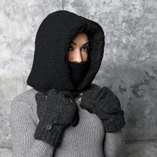 A person wearing the black Hand Knit Glide Hood, a matching face covering, gray gloves, and a ribbed gray sweater stands against a textured gray background, with only their eyes visible.