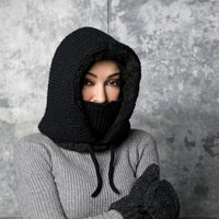 A person wears the Hand Knit Glide Hood—a black, hand-knit lambswool hood with a face covering—along with a gray ribbed sweater and dark gray knit mittens, standing against a textured gray background and gazing directly at the camera.