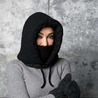 A person wears the Hand Knit Glide Hood—a black, hand-knit lambswool hood with a face covering—along with a gray ribbed sweater and dark gray knit mittens, standing against a textured gray background and gazing directly at the camera.