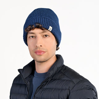 A person wears the Baltic Hat, a blue wool hat handmade in Nepal, along with a black quilted jacket and a blue crew neck shirt, standing against a plain white background.