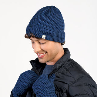 A person in the Baltic Hat, a blue wool beanie handmade in Nepal, paired with matching knit mittens and a black puffer jacket, smiles while looking down against a plain white background.