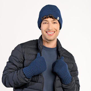 Smiling at the camera with both thumbs up, a person wears a dark blue knit hat, a blue sweater, a dark puffer jacket, and Kent Mittens. The background is plain white.