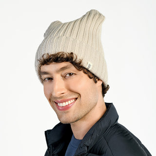 A man with curly hair is smiling and wearing the Ely Hat, a beige wool beanie with faux sherling lining, and a dark jacket against a plain white background.