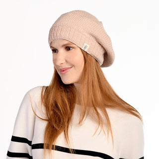 A woman with long red hair wears the Abbey Hat, a light pink wool beanie handmade in Nepal, along with a cream sweater featuring black horizontal stripes. She stands slightly turned and smiling against a plain white background.
