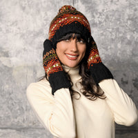 A person in a cream turtleneck sweater poses in front of a textured gray wall, wearing a matching patterned knit hat and the Hand Knit Explore Mittens, which feature Fair Isle designs in red, orange, yellow, and black colors, while smiling at the camera.