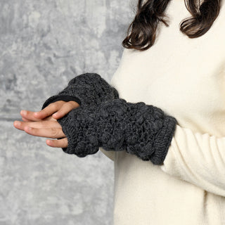 Wearing a cream sweater, a person displays the Flower Crochet Handwarmers—dark gray, chunky knit fingerless gloves ideal for keeping hands warm. Their hands are held together against a gray textured background, with only their torso and arms visible.