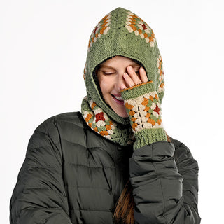 Wearing Ryan Handwarmers with multicolored patterns and a green crochet hood, a smiling person in a dark puffer jacket covers part of their face and stands against a plain background.