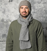 A man with a beard wears a gray hand-crocheted hat and a cozy Hand Crochet Velour Scarf, paired with an olive green jacket. He is standing in front of a textured gray background, facing the camera with a neutral expression.