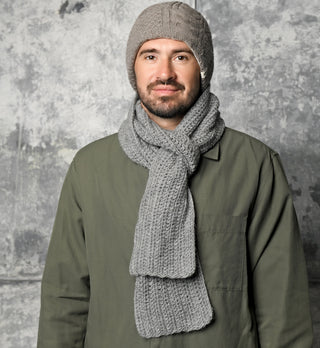 A man with a beard wears a gray hand-crocheted hat and a cozy Hand Crochet Velour Scarf, paired with an olive green jacket. He is standing in front of a textured gray background, facing the camera with a neutral expression.