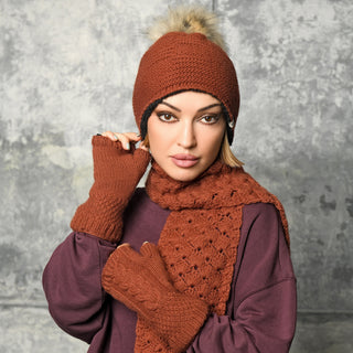 Wearing rust-colored Cable Handwarmers, along with a matching knit hat featuring a pom-pom and scarf, a woman poses confidently against a textured gray background. She also sports a purple sweater and gazes directly at the camera.