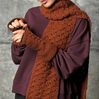 A person wearing a purple sweatshirt, a hand crocheted brown Merino wool Hand Crochet Veil Scarf featuring a textured bobble stitch, and matching fingerless gloves stands against a gray textured background. Only the lower half of their face is visible.