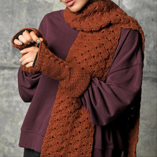 A person wearing a purple sweatshirt, a hand crocheted brown Merino wool Hand Crochet Veil Scarf featuring a textured bobble stitch, and matching fingerless gloves stands against a gray textured background. Only the lower half of their face is visible.