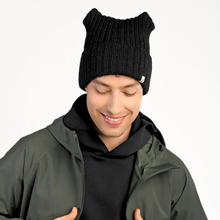 A person wears the Ely Hat, a handmade black knit beanie with cat ear shapes from Nepal, along with a black hoodie and dark green jacket, smiling and looking down against a plain white background.