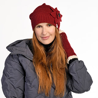 A woman with long red hair wears a gray puffer jacket, the Monroe Hat—a handmade in Nepal red wool beanie with a flower detail—and matching fingerless gloves with white trim. She smiles slightly and holds one hand to her head against a white background.