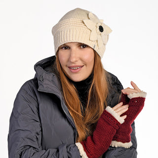 A woman with long reddish-brown hair poses against a white background wearing a gray puffy jacket, red fingerless gloves with cream trim, and the Monroe Hat—a wool beanie handmade in Nepal.