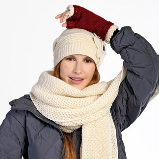 Wearing the Ithaca Scarf in cream with a matching hat, gray puffy jacket, and red fingerless glove, a person poses with one arm bent and hand on their head, smiling at the camera—a stylish and cozy winter accessory look.
