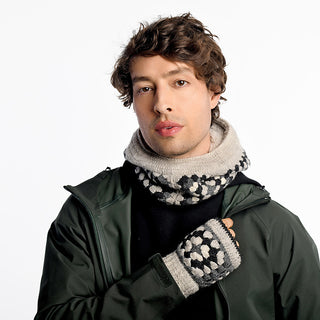 A person with wavy brown hair wears a green jacket, black shirt, beige patterned scarf, and Ryan Handwarmers. The background is plain white.