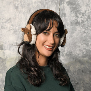 A woman with wavy dark hair smiles at the camera, wearing a dark green sweater and the Hand Crochet Grizzly Earmuffs—brown, bear-themed earmuffs made from 100% wool with an adjustable headband. The background is a textured gray wall.