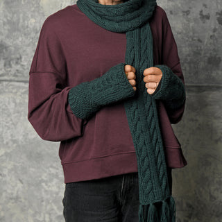 A person wearing a maroon sweatshirt, dark pants, and the Hand Knit Plush Scarf in dark green with matching fingerless gloves stands in front of a textured gray wall. The scarf is crafted from soft Merino wool, features fringe ends, and their face is not visible.