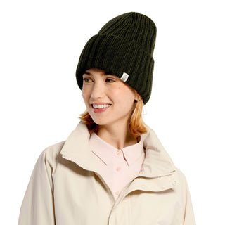 A person with light skin and short, light brown hair wears the Cooper Hat—a dark ribbed merino wool winter hat—paired with a pale collared shirt and a light beige jacket. They are looking slightly to the side and smiling.